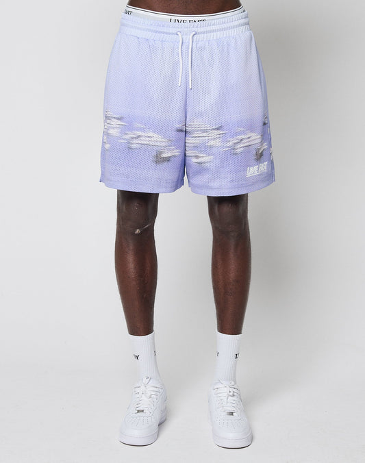 A person wears LFDY Boat Mesh Shorts by LIVE FAST DIE YOUNG in light purple with a cloud pattern and “LIVE FAST” logo on the lower left leg, styled with white socks and sneakers, standing against a plain white background. Made from soft polyester.
