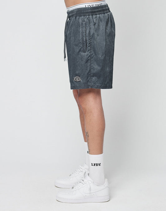 A person stands sideways in LIVE FAST DIE YOUNG Live Fast Swimshorts, featuring a visible elastic waistband with LIVE FAST text, paired with LFDY white socks and white sneakers, against a plain white background.