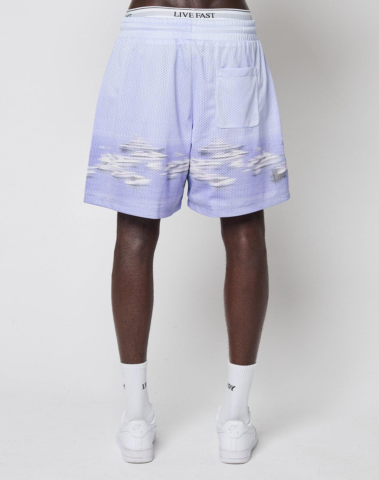 A person stands facing away, wearing LIVE FAST DIE YOUNG LFDY Boat Mesh Shorts in light purple with a cloud pattern, white LIVE FAST waistband, and a back pocket—paired with white socks and sneakers.