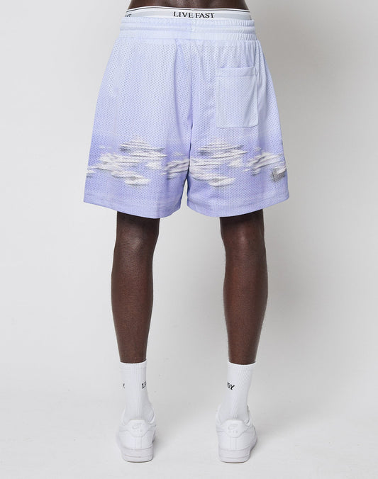 A person stands facing away, wearing LIVE FAST DIE YOUNG LFDY Boat Mesh Shorts in light purple with a cloud pattern, white LIVE FAST waistband, and a back pocket—paired with white socks and sneakers.