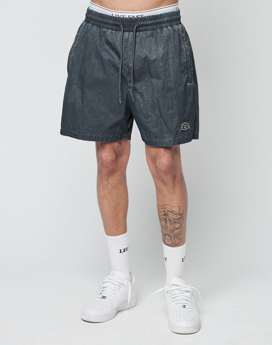 A person wears LIVE FAST DIE YOUNG Live Fast Swimshorts in dark gray with an elastic waistband and drawstring, white LE FIX crew socks, white sneakers, and has a tattoo on their left leg, standing against a light background.