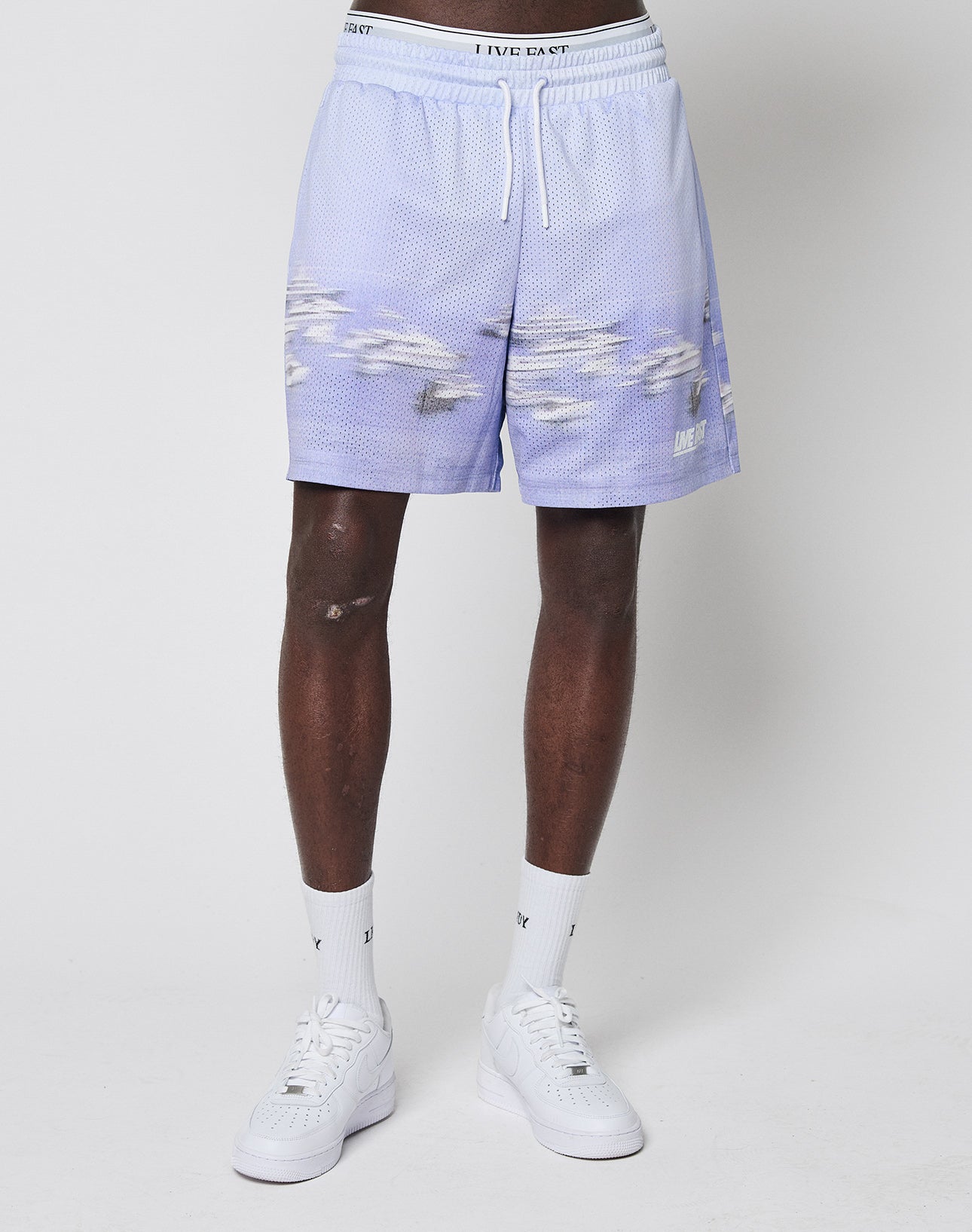 A person wears LIVE FAST DIE YOUNG LFDY Boat Mesh Shorts in light purple with a white fish pattern, paired with white socks and sneakers, standing against a light gray background. Only the lower half is visible. Polyester fabric ensures comfort and style.