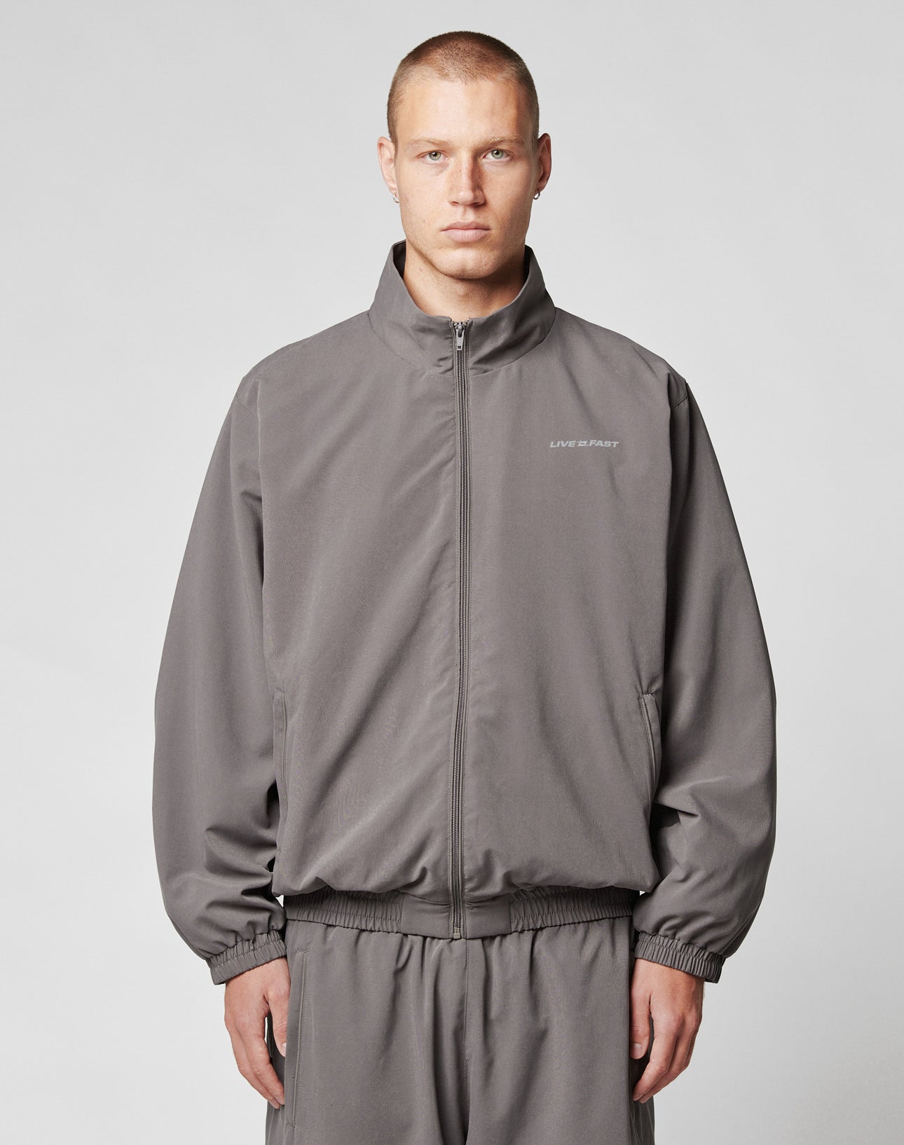 A young man with short hair wears the LIVE FAST DIE YOUNG LFDY Trackjacket and matching polyester pants in gray, standing against a plain light gray background and looking at the camera with a neutral expression.