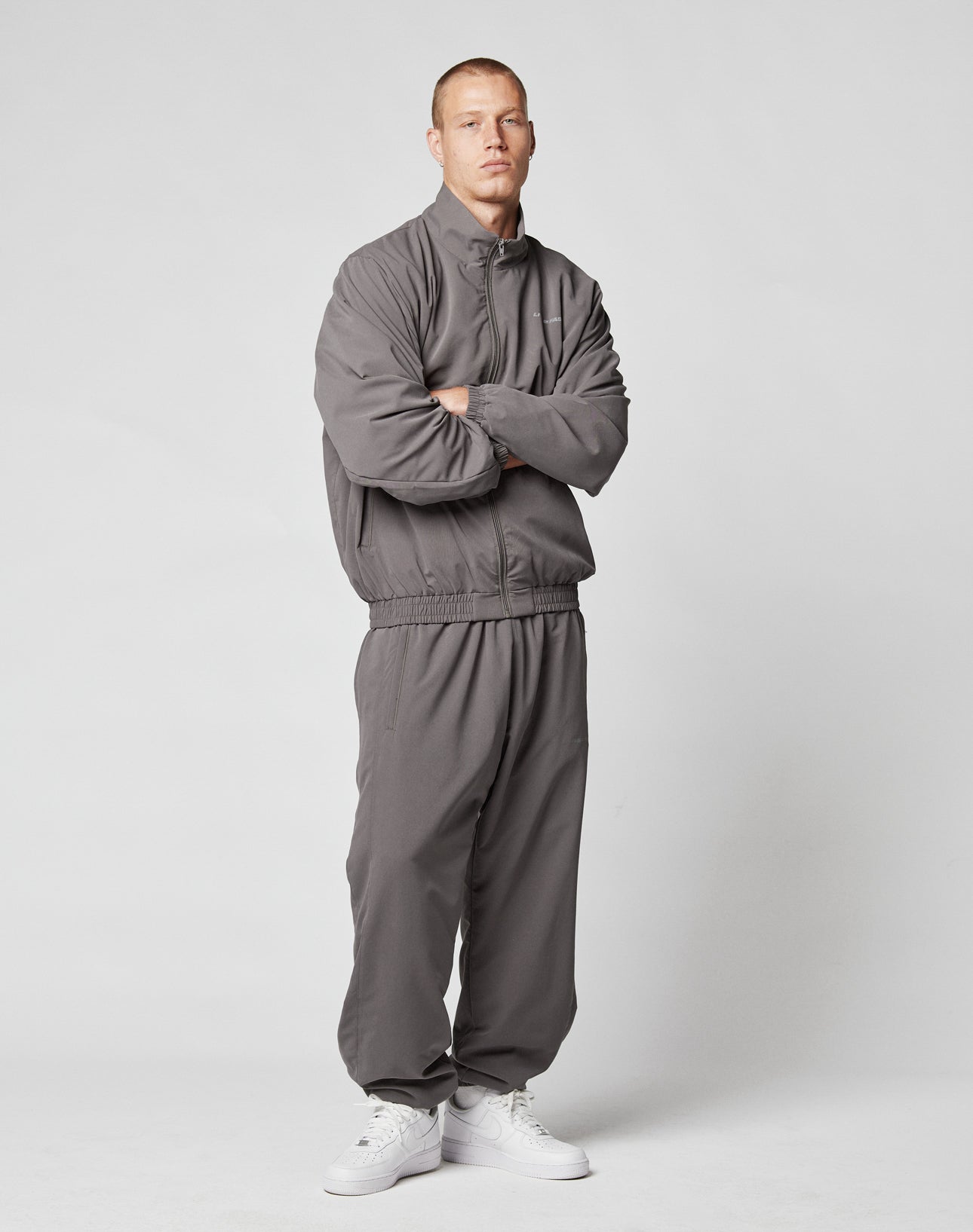 A man with arms crossed wears LIVE FAST DIE YOUNG LFDY Trackpants in gray (regular fit, Elastikbund) and white sneakers, standing against a plain light gray background.