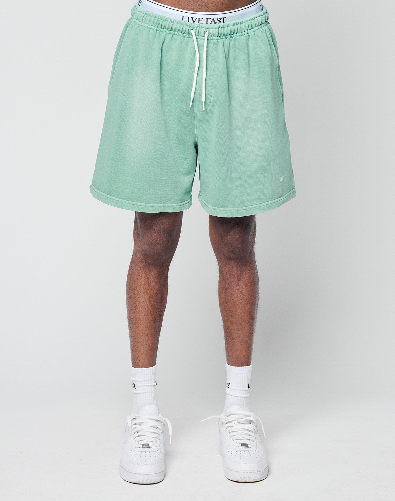 A person stands against a plain background wearing LIVE FAST DIE YOUNG Basic Sweat Shorts in light green with individueller Spray Effekt, white socks, white sneakers, and visible white LIVE FAST waistband underwear above the Regular Fit shorts.