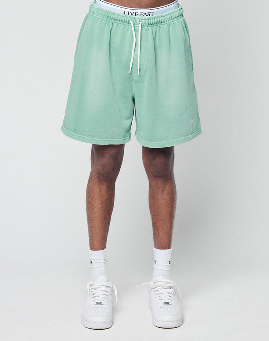A person stands against a plain background wearing LIVE FAST DIE YOUNG Basic Sweat Shorts in light green with individueller Spray Effekt, white socks, white sneakers, and visible white LIVE FAST waistband underwear above the Regular Fit shorts.