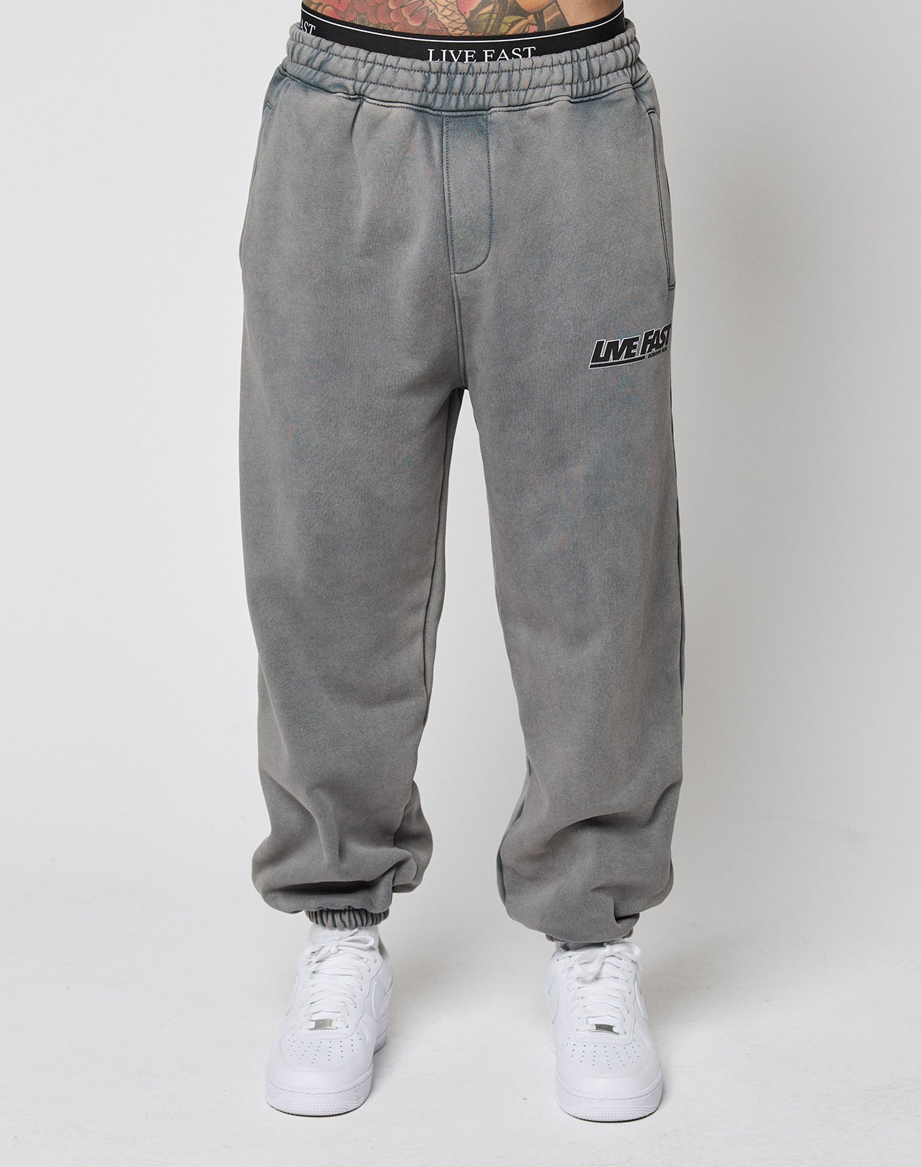 A person wears the LIVE FAST DIE YOUNG LFDY Europe Run Jogger—loose-fitting, faded gray sweatpants with LIVE FAST on the left thigh, Vintage Waschung, and elasticized waistband and cuffs—paired with white sneakers against a white background.
