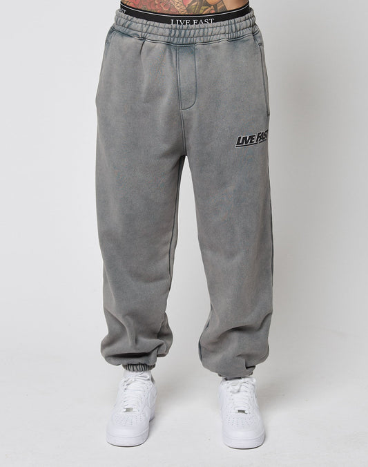 A person wears the LIVE FAST DIE YOUNG LFDY Europe Run Jogger—loose-fitting, faded gray sweatpants with LIVE FAST on the left thigh, Vintage Waschung, and elasticized waistband and cuffs—paired with white sneakers against a white background.