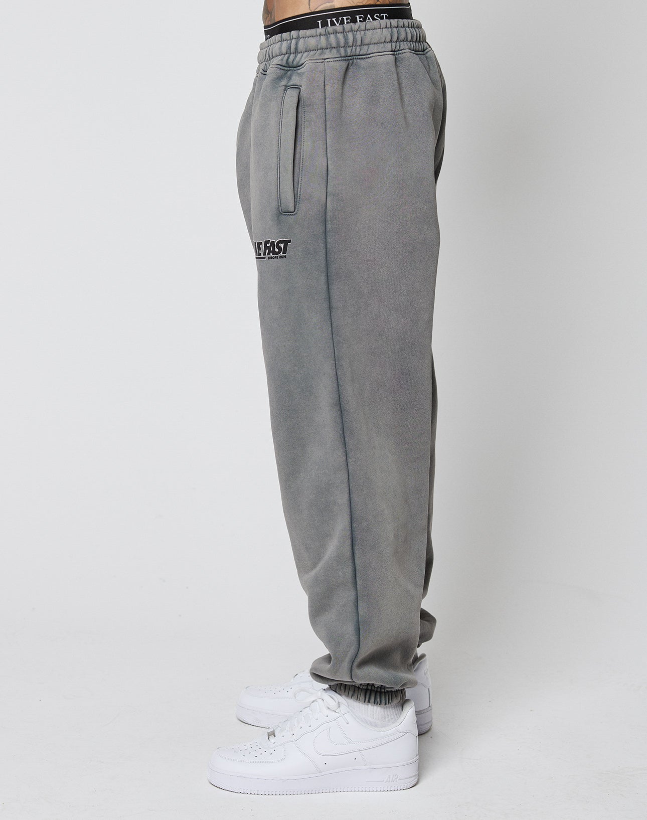 A person wears the LFDY Europe Run Jogger by LIVE FAST DIE YOUNG, featuring a vintage wash, elastic waistband with LIVE FAST text, side pockets, and white sneakers. Side view photo against a plain white background.