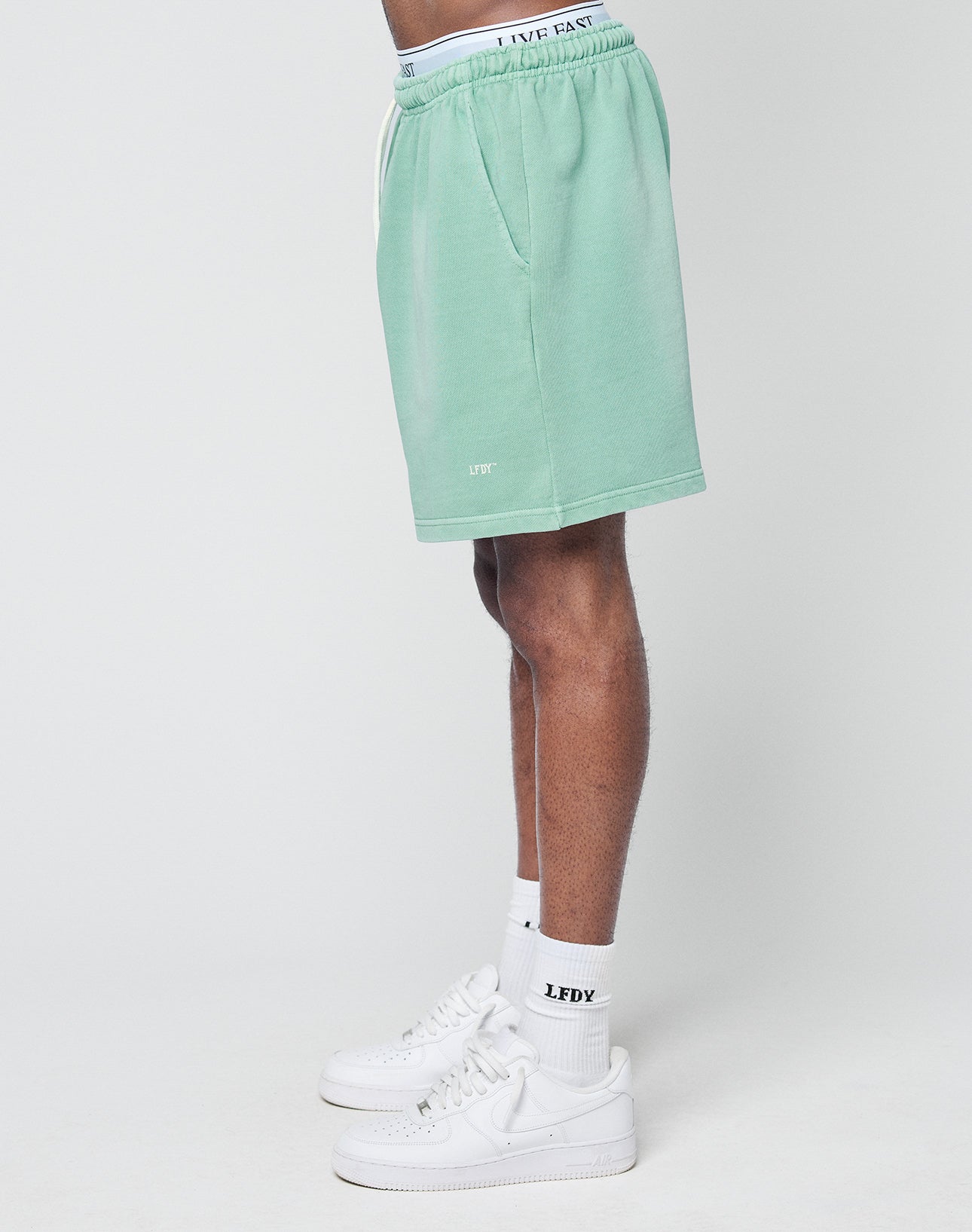 A person stands sideways against a plain light background wearing LIVE FAST DIE YOUNG Basic Sweat Shorts in mint green with handmade fade effect, featuring LIVE FAST on the waistband, paired with white LFV socks and sneakers.