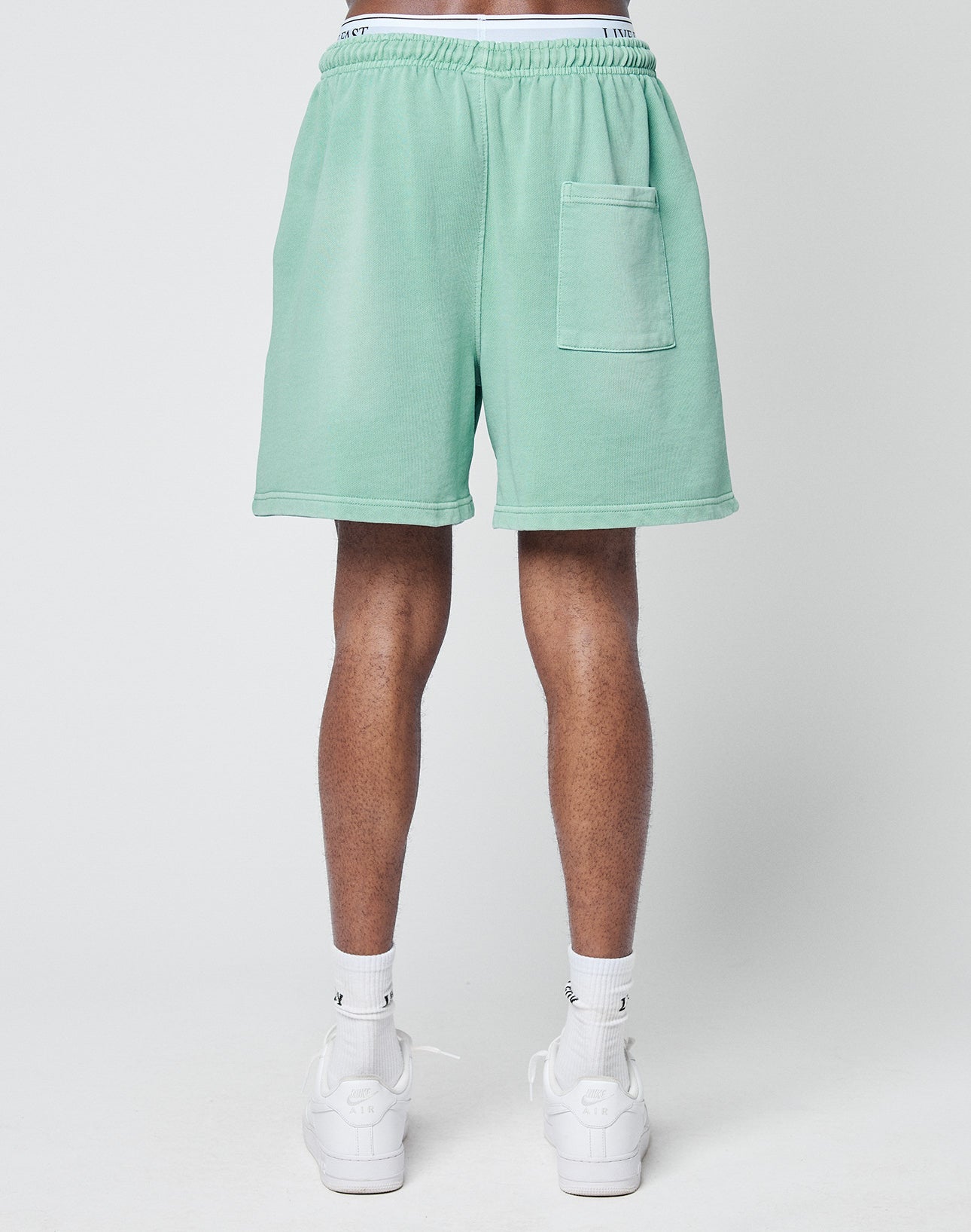 A person faces away, wearing LIVE FAST DIE YOUNG Basic Sweat Shorts in light green with a back pocket, white waistband, white crew socks, and white sneakers against a plain light background.