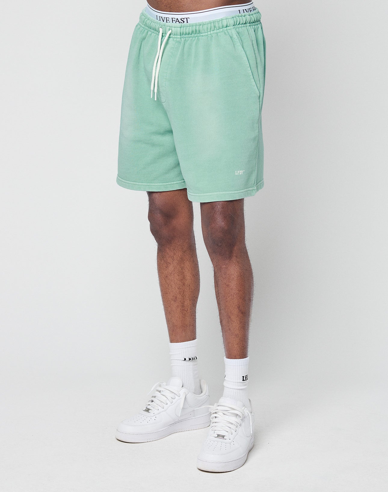 A person models light green Basic Sweat Shorts by LIVE FAST DIE YOUNG, featuring a regular fit, drawstring waist, and LIVE text near the hem. Only the lower half is shown, styled with white socks and sneakers against a plain white background.
