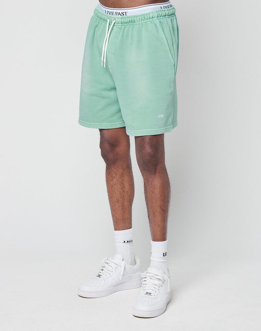 A person models light green Basic Sweat Shorts by LIVE FAST DIE YOUNG, featuring a regular fit, drawstring waist, and LIVE text near the hem. Only the lower half is shown, styled with white socks and sneakers against a plain white background.