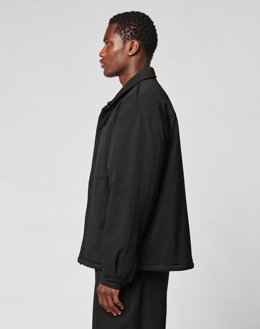 A person with short curly hair stands in profile, wearing the LIVE FAST DIE YOUNG Padded Casual Shirt and oversized black pants against a plain light gray background.