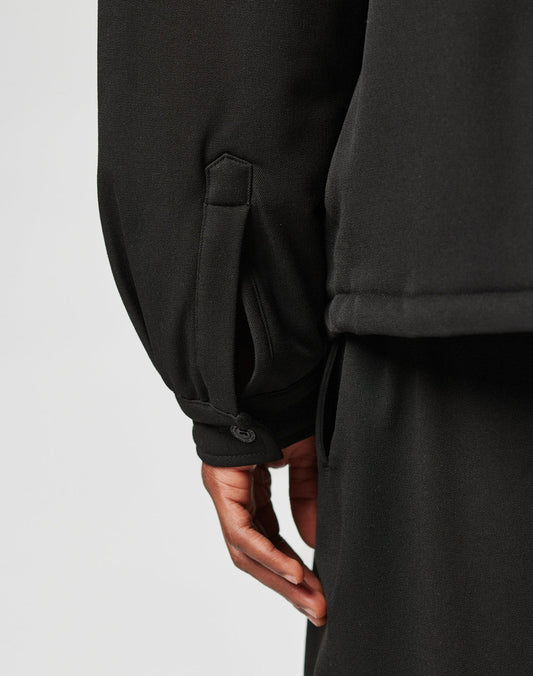 Close-up of a person’s hand at their side, wearing LIVE FAST DIE YOUNG’s Padded Casual Shirt with a buttoned strap detail at the cuff and matching black pants. The background is plain and light-colored.