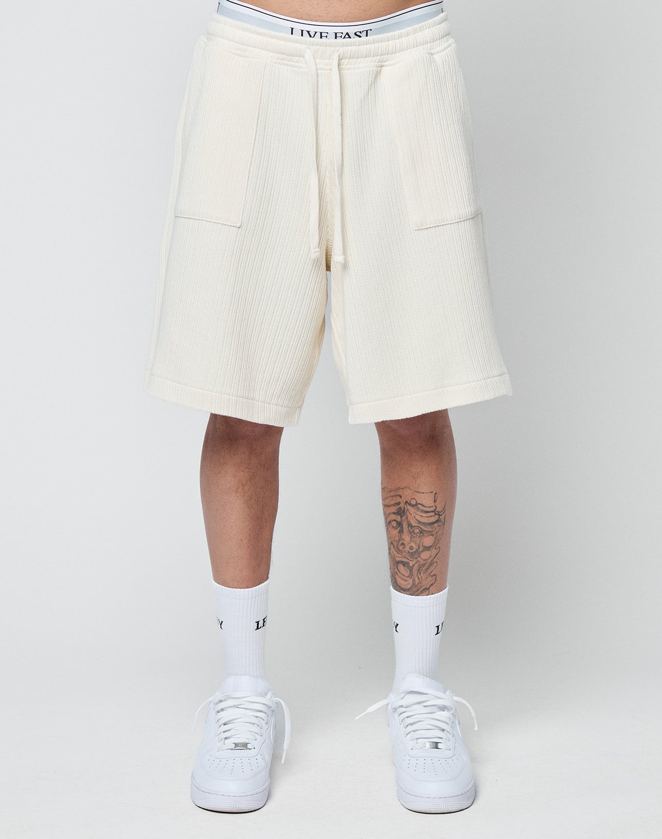 Someone wears LIVE FAST DIE YOUNG Basic Knit Shorts in cream with an elastic waistband and patch pockets, paired with white socks and sneakers. A tattoo is partly visible on their left leg against a plain light gray background.