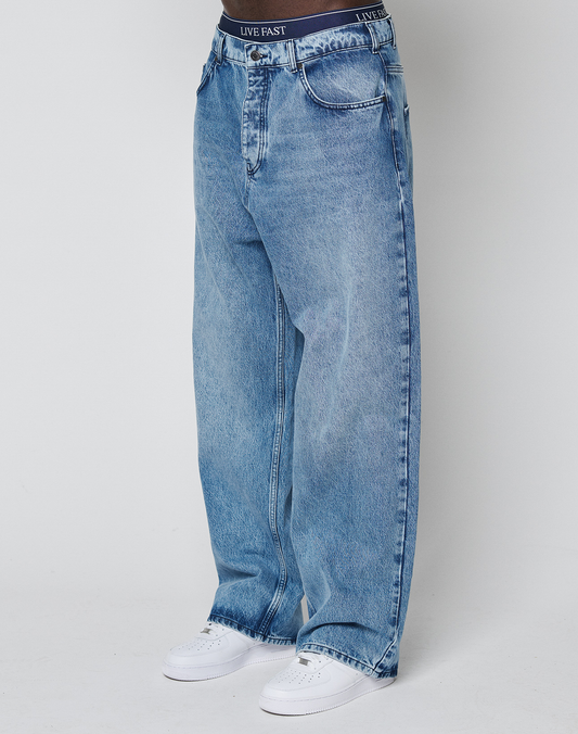 A person wears the LFDY Baggy Denim by LIVE FAST DIE YOUNG—baggy, light blue jeans with a wide straight leg and baggy fit, made of 100% cotton—paired with a black LIVE FAST waistband and white sneakers against a white background. | model
