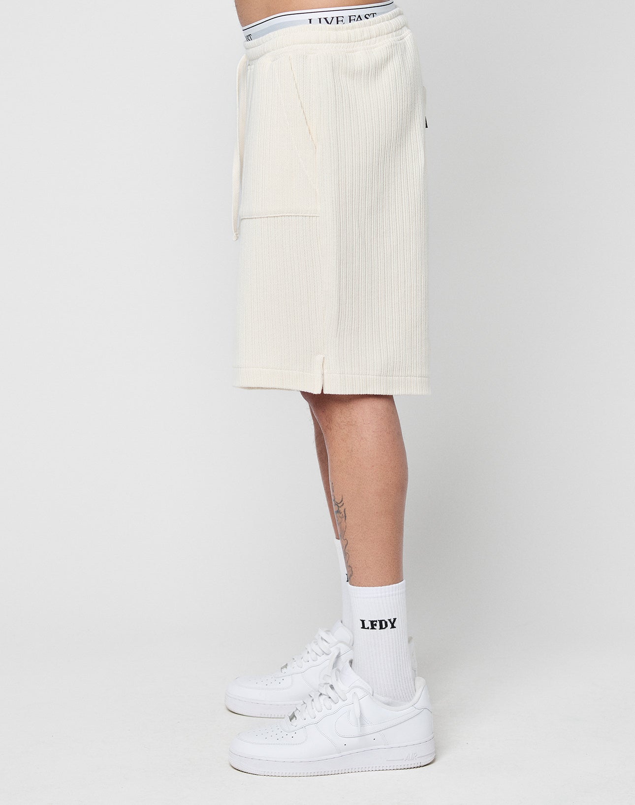 A person stands sideways against a plain white background, wearing LIVE FAST DIE YOUNG Basic Knit Shorts in cream with patch pockets and an elastic waistband, plus white LFDY socks, Nike sneakers, and a visible LIVE waistband.