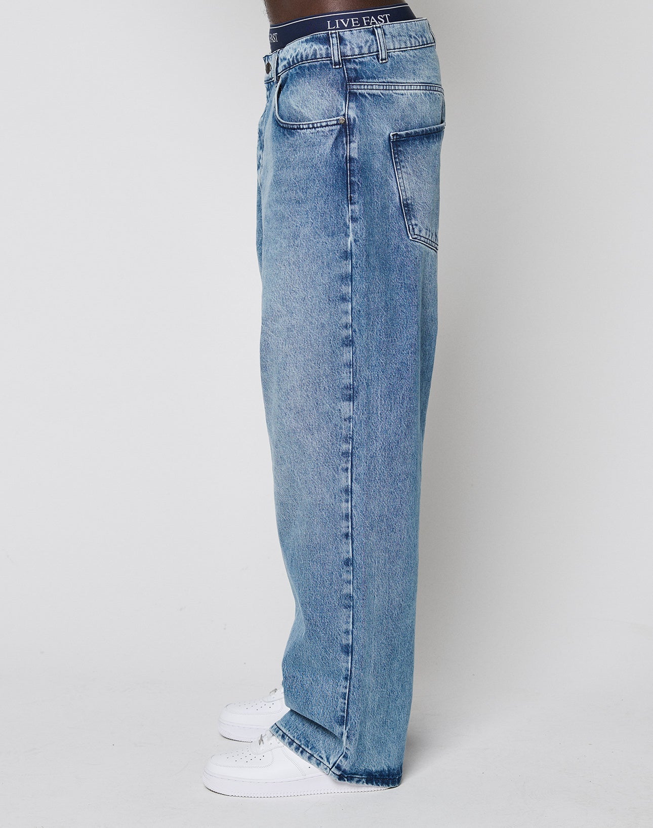 A person wearing the LFDY Baggy Denim by LIVE FAST DIE YOUNG—loose-fitting blue jeans with a wide straight leg made from 100% cotton—and white sneakers stands sideways against a plain white background, showing only the lower half.