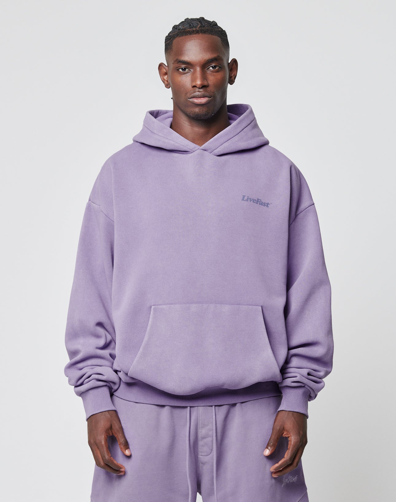 A man faces forward in an oversize, light purple Basic 360 Hooded and matching sweatpants by LIVE FAST DIE YOUNG, both featuring LiveFast text, made from Baumwolle Polyester, against a plain white background.