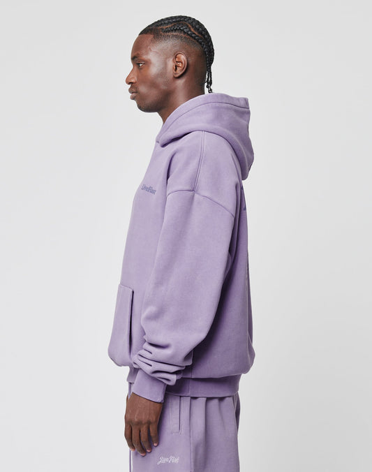 A man with braided hair stands in profile, wearing the Basic 360 Hooded and matching sweatpants by LIVE FAST DIE YOUNG. This oversize fit lavender hoodie is made from Baumwolle Polyester, with a front pocket and subtle chest text on white.
