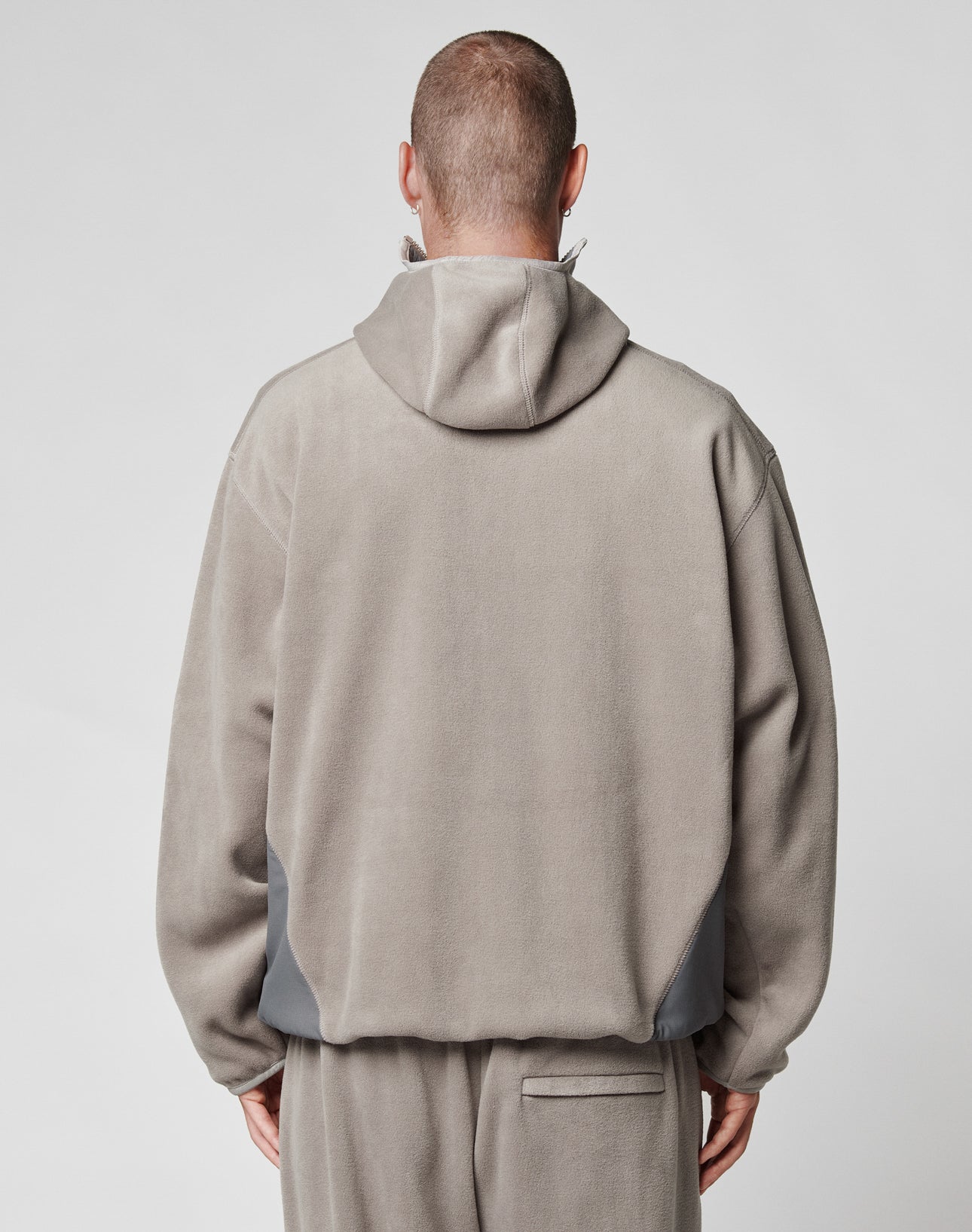 A person is seen from behind wearing the LIVE FAST DIE YOUNG Live Fast Fleece Halfzip in light gray with matching pants. The oversize fit jacket features a subtle nylon einsatz detail, hood up, head shaved, set against a plain background.
