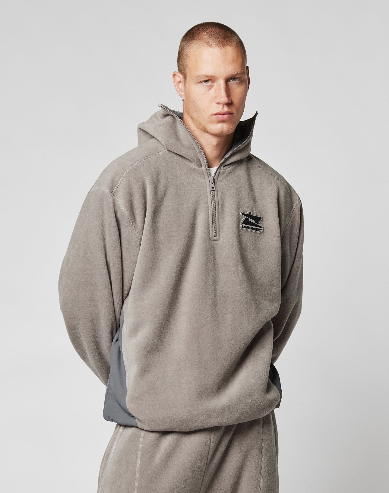 Someone with short hair wears the LIVE FAST DIE YOUNG Live Fast Fleece Halfzip, a loose, light gray, oversized fleece pullover with a half-zip and small black chest logo, standing against a plain light background.