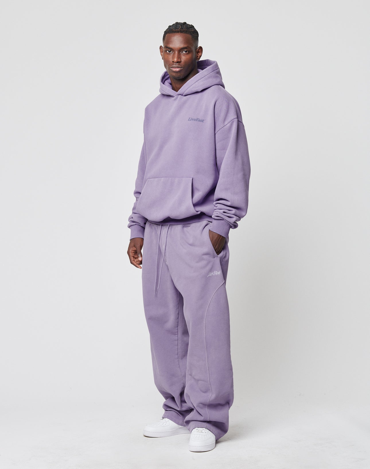 A man poses against a plain background wearing the LIVE FAST DIE YOUNG Basic Wide Jogger in lavender with matching hoodie. He pairs the wide fit set with white sneakers, smiles at the camera, and keeps one hand in his pocket.