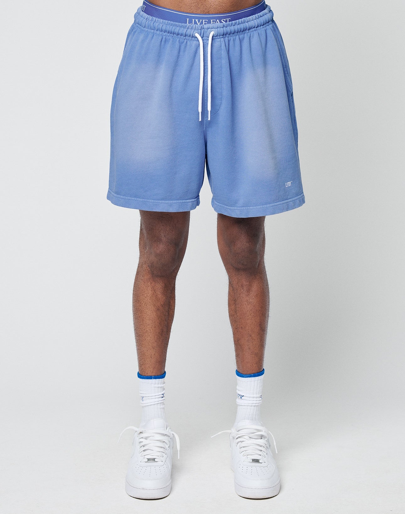 A person stands against a light gray background, wearing LIVE FAST DIE YOUNG Basic Sweat Shorts in light blue with an elastic waistband, paired with white socks and sneakers. The image is cropped from the waist down.