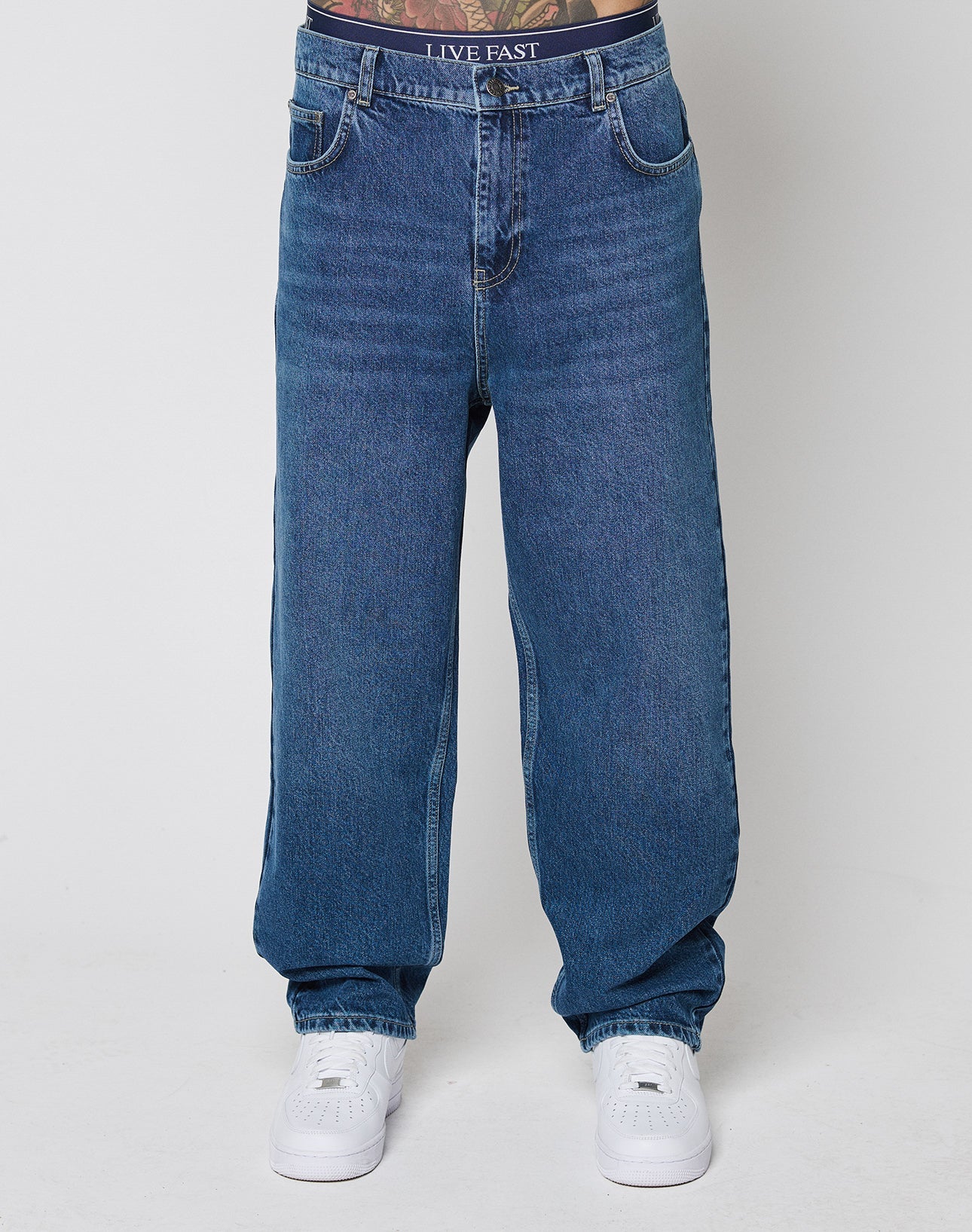 A model in LIVE FAST DIE YOUNG Wide Denim blue jeans (regular fit, wide leg, 100% Baumwolle) and white sneakers shows a black waistband labeled LIVE FAST above the jeans. Plain white background.