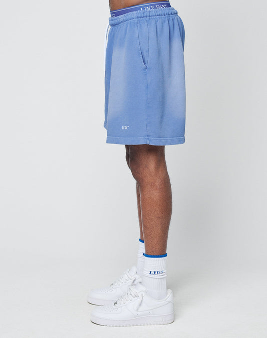 A person stands sideways, showing only the lower body, wearing LIVE FAST DIE YOUNG Basic Sweat Shorts in light blue with a faded look and elastic waistband, paired with white crew socks with blue accents and white sneakers against a plain background.