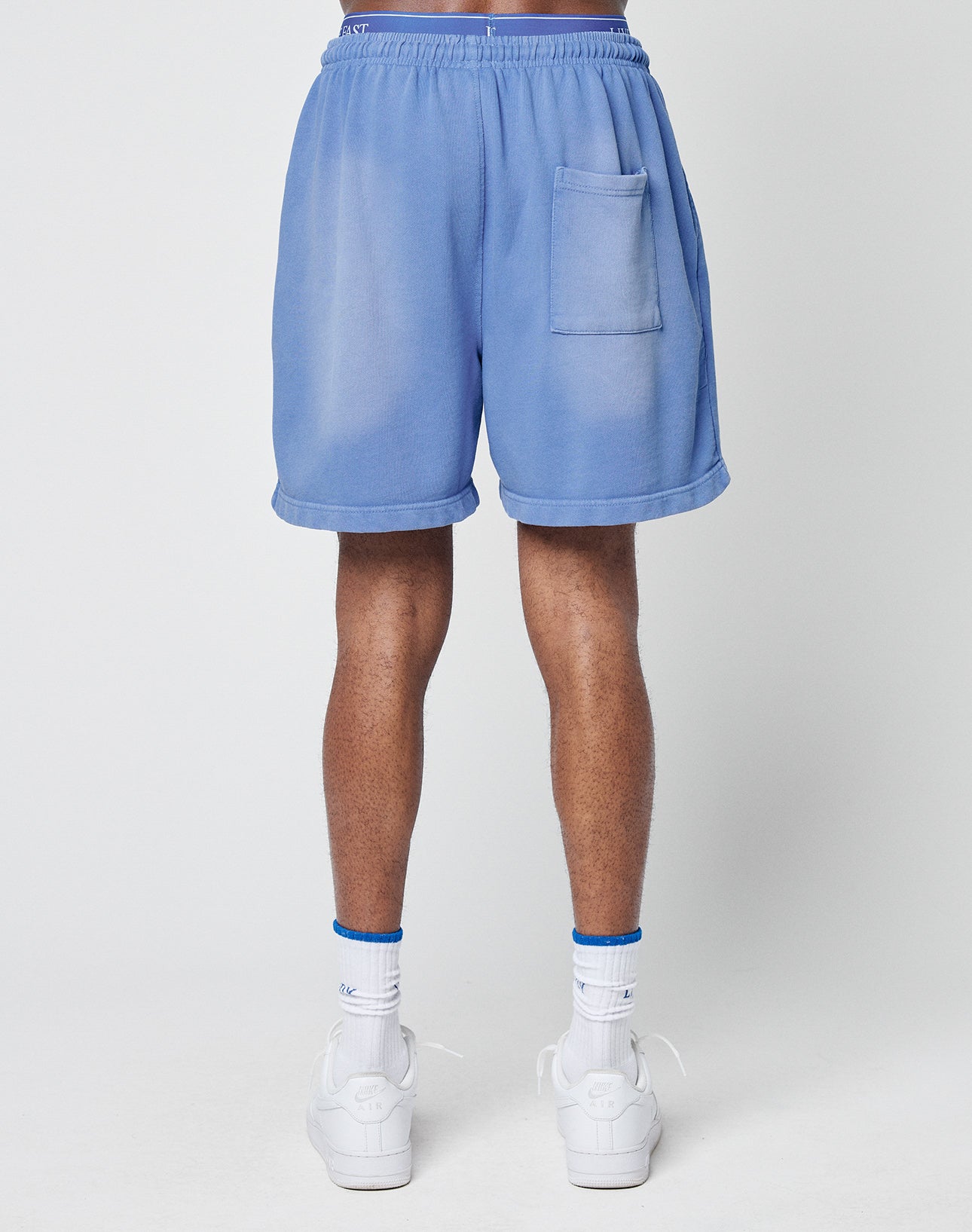 A person stands facing away, wearing LIVE FAST DIE YOUNG Basic Sweat Shorts in light blue with a faded look and back pocket, paired with white socks and sneakers. The background is plain and light-colored.