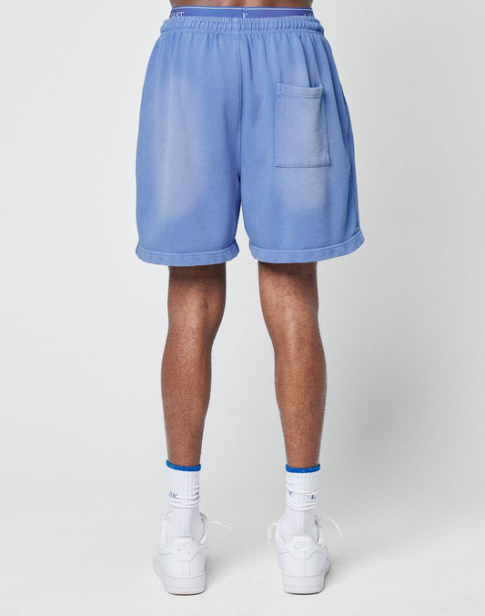 A person stands facing away, wearing LIVE FAST DIE YOUNG Basic Sweat Shorts in light blue with a faded look and back pocket, paired with white socks and sneakers. The background is plain and light-colored.