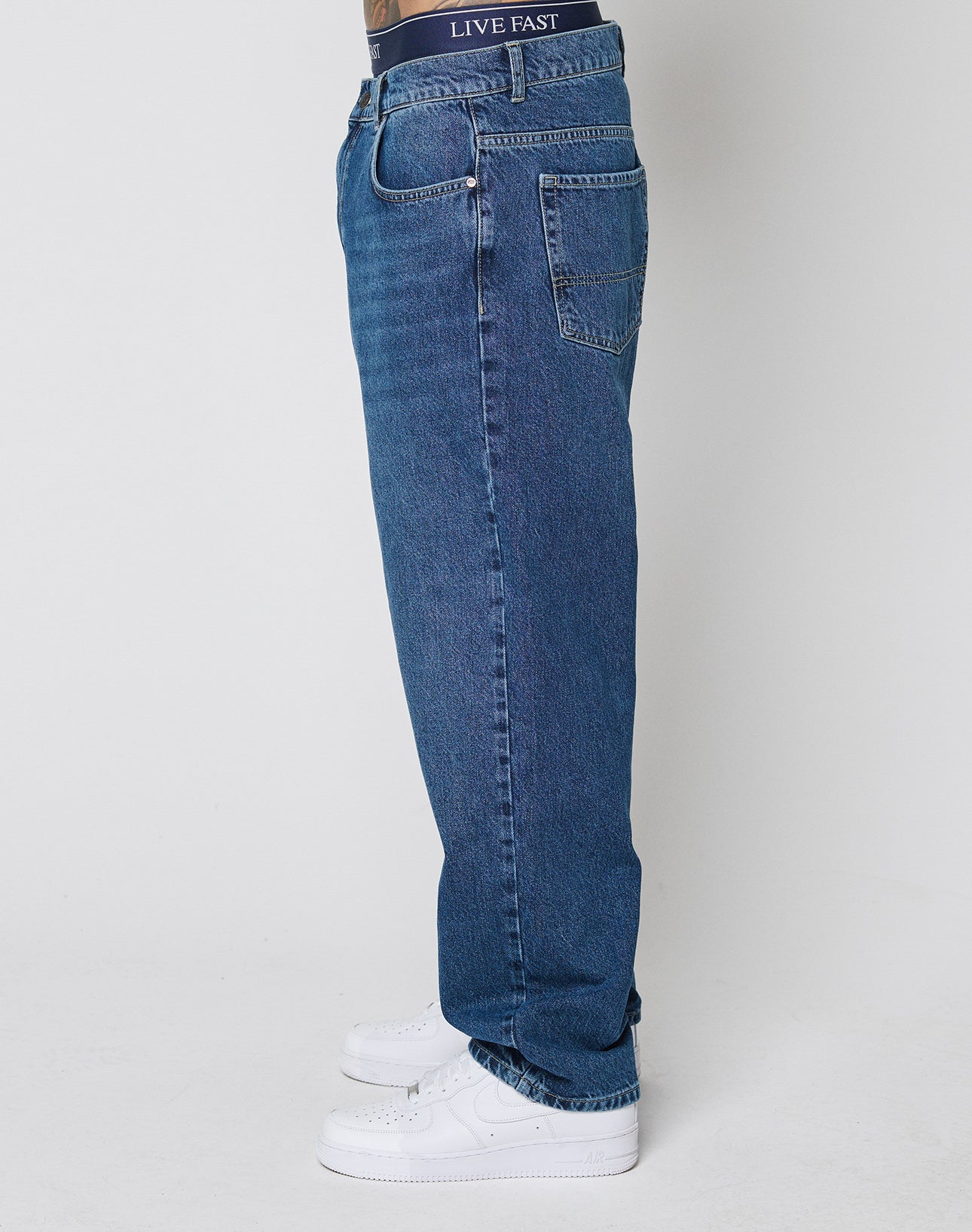 A person stands sideways in LIVE FAST DIE YOUNG Wide Denim, loose-fitting blue jeans with LIVE FAST on the waistband and white sneakers. The wide-leg jeans are 100% Baumwolle. The background is plain white.