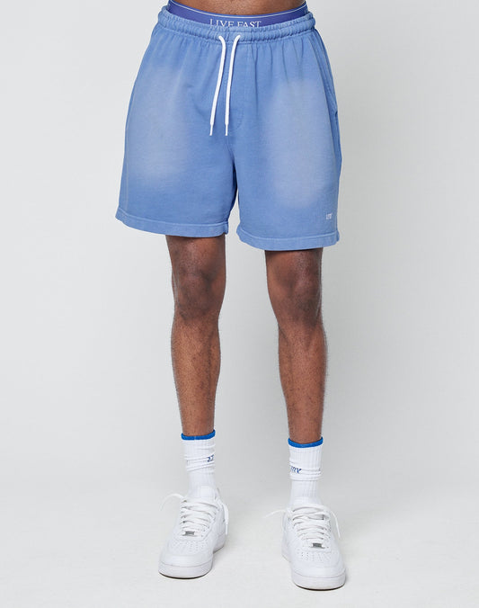 A person stands facing forward against a white background, wearing LIVE FAST DIE YOUNG Basic Sweat Shorts in light blue with a faded look, white drawstring and elastic waistband, paired with white socks and sneakers.