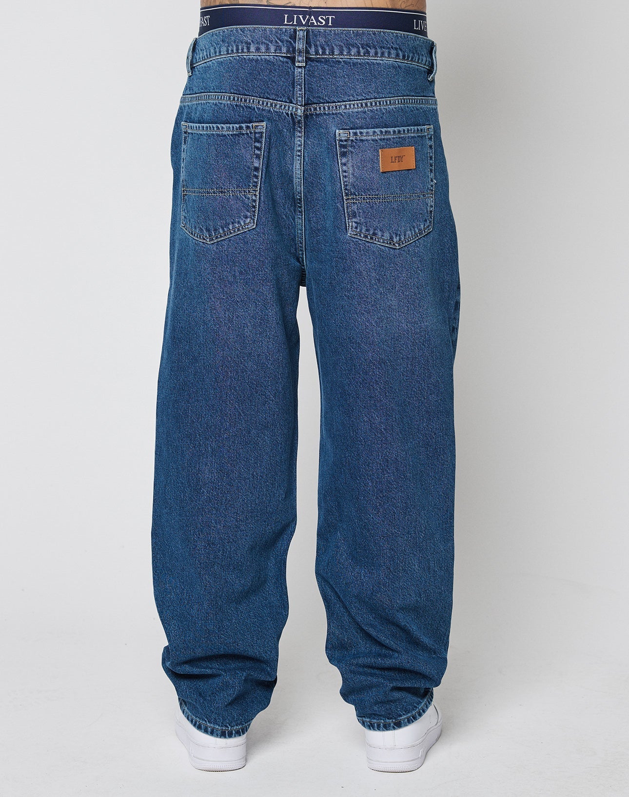 A person seen from the back wears LIVE FAST DIE YOUNG Wide Denim jeans with visible dark underwear above the waistband, two back pockets, a brown leather patch on one pocket, and white shoes.