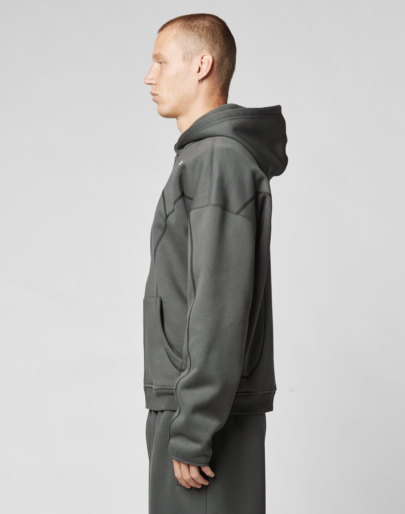 A person with short hair stands in profile, facing left, wearing the LIVE FAST DIE YOUNG LFDY Active Fleece Hooded Zip and matching pants in dark grey cotton, set against a plain light grey background.