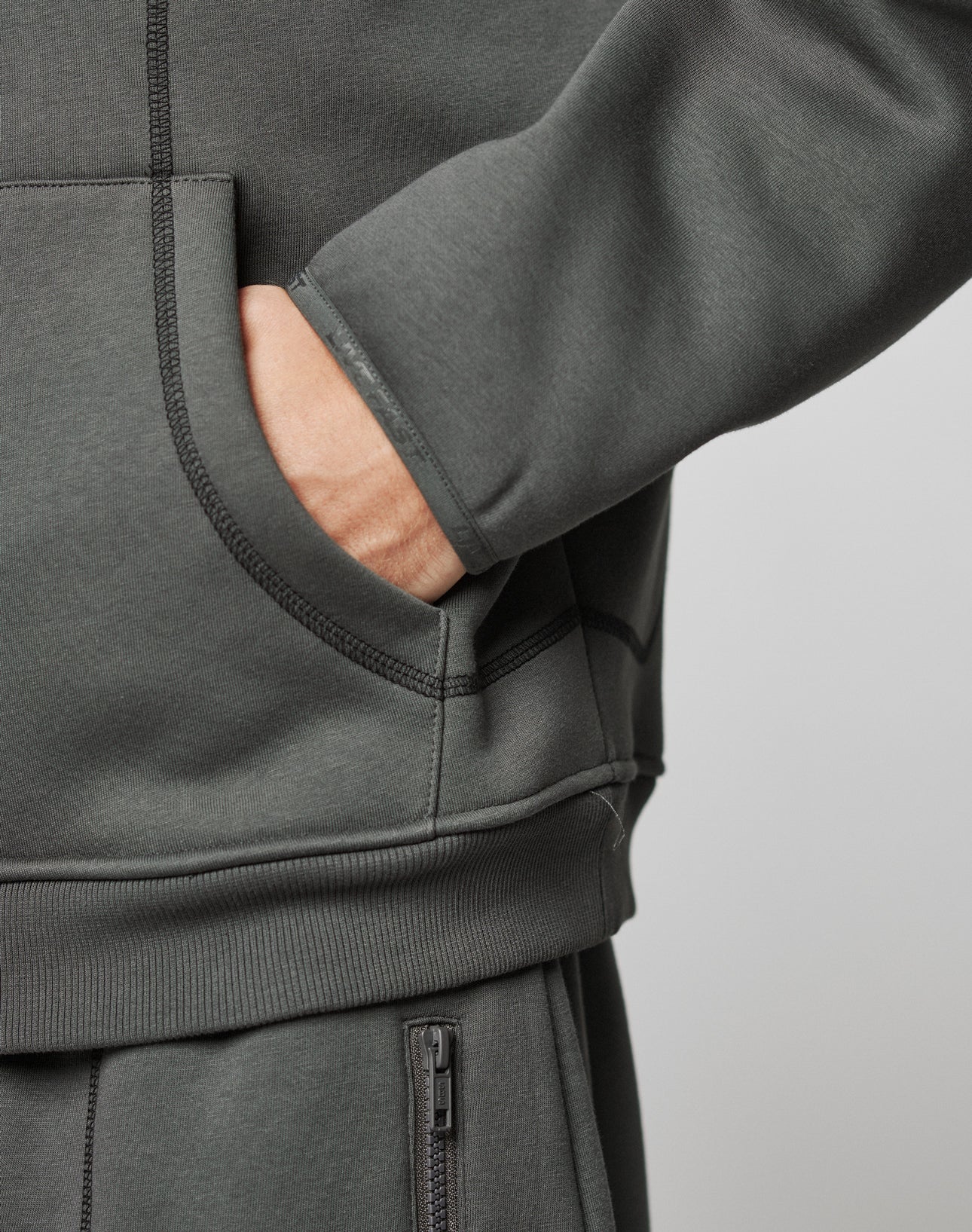 Wearing the LFDY Active Fleece Hooded Zip by LIVE FAST DIE YOUNG in dark gray, a person sports an oversize fit with matching sweatpants, zippered pocket, visible stitching, subtle logo print details, and right hand in the front pocket.