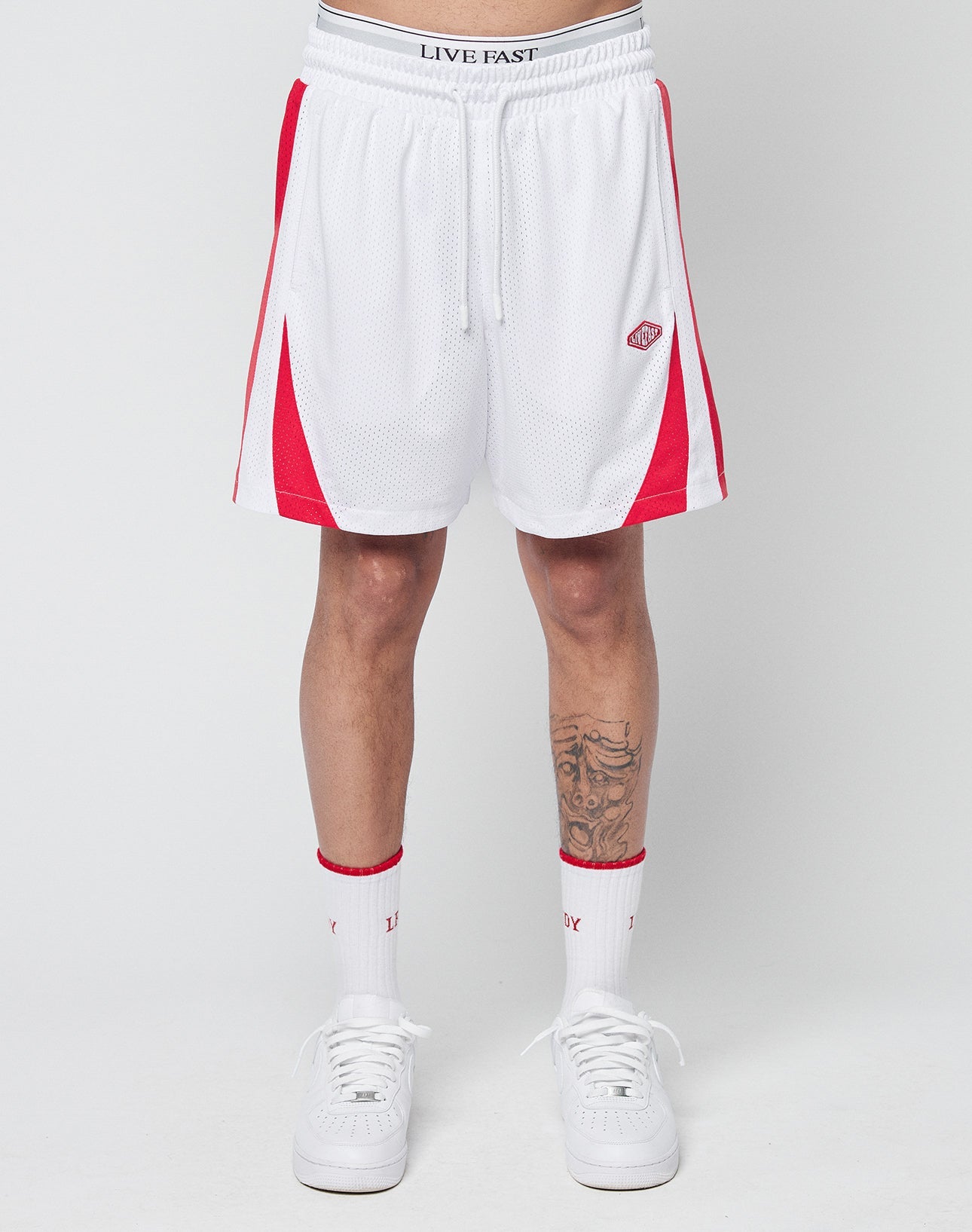 A person wearing LIVE FAST DIE YOUNG Striped Mesh Shorts in white with red side panels, paired with white socks with red accents and white sneakers. Only their legs and lower torso are visible. The shorts have a logo patch on the left leg.