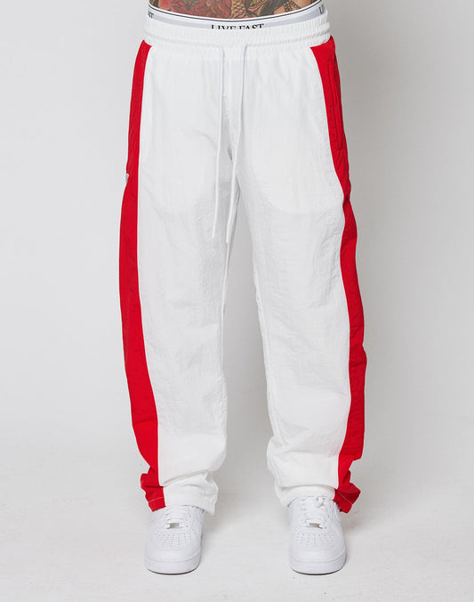 A person wearing LIVE FAST DIE YOUNG LFDY Trackpants 3000—wide fit, white with bold red side stripes, elastic waistband, and crafted from lightweight polyamide—pairs them with white sneakers against a plain white background.
