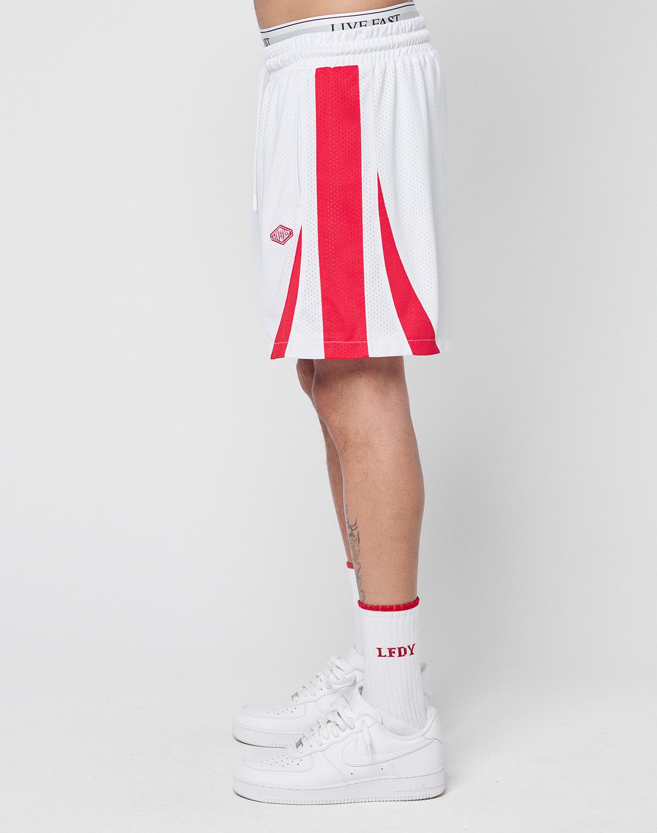 A person stands sideways against a plain white background wearing LIVE FAST DIE YOUNG Striped Mesh Shorts, featuring a regular fit with white fabric, red vertical stripes, and a logo patch, paired with white crew socks and white Nike sneakers.