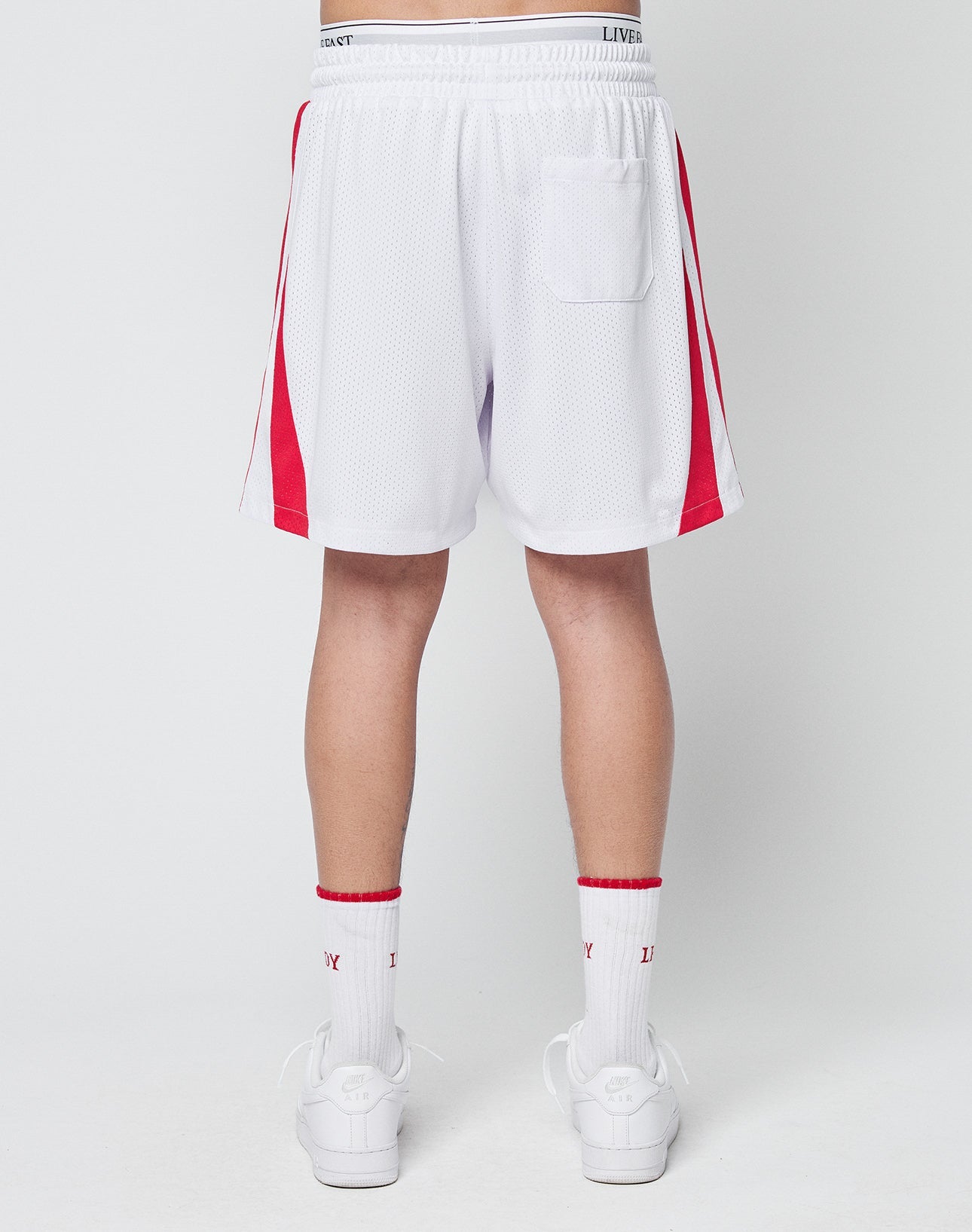 A person faces away, wearing LIVE FAST DIE YOUNG Striped Mesh Shorts—white polyester with red side stripes, a logo patch, and a single right back pocket—paired with white socks featuring red accents and white sneakers.