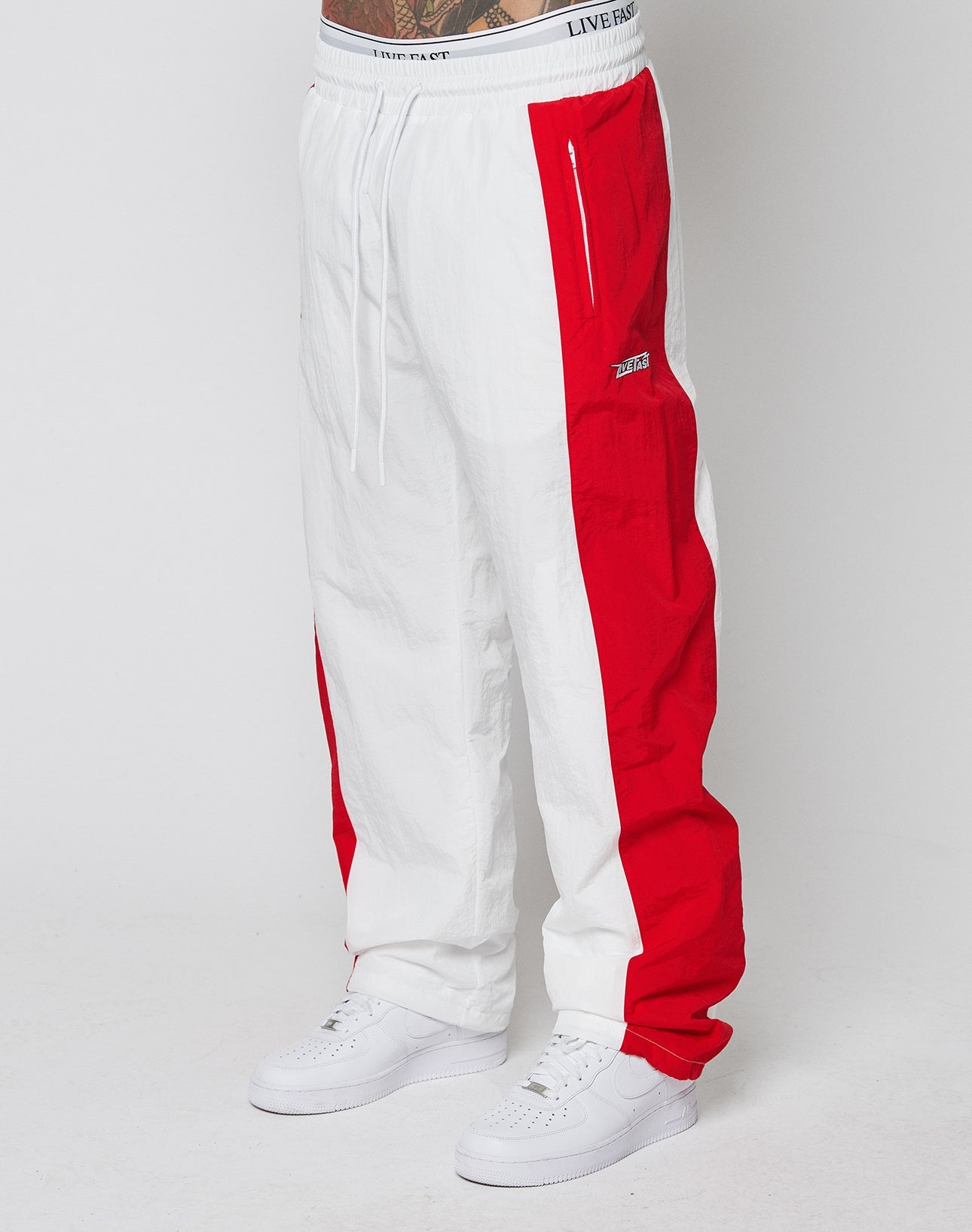 A person wears LFDY Trackpants 3000 by LIVE FAST DIE YOUNG—wide fit, white and red color-blocked with elastic waistband and zippered pockets—paired with white sneakers, standing against a plain white background.