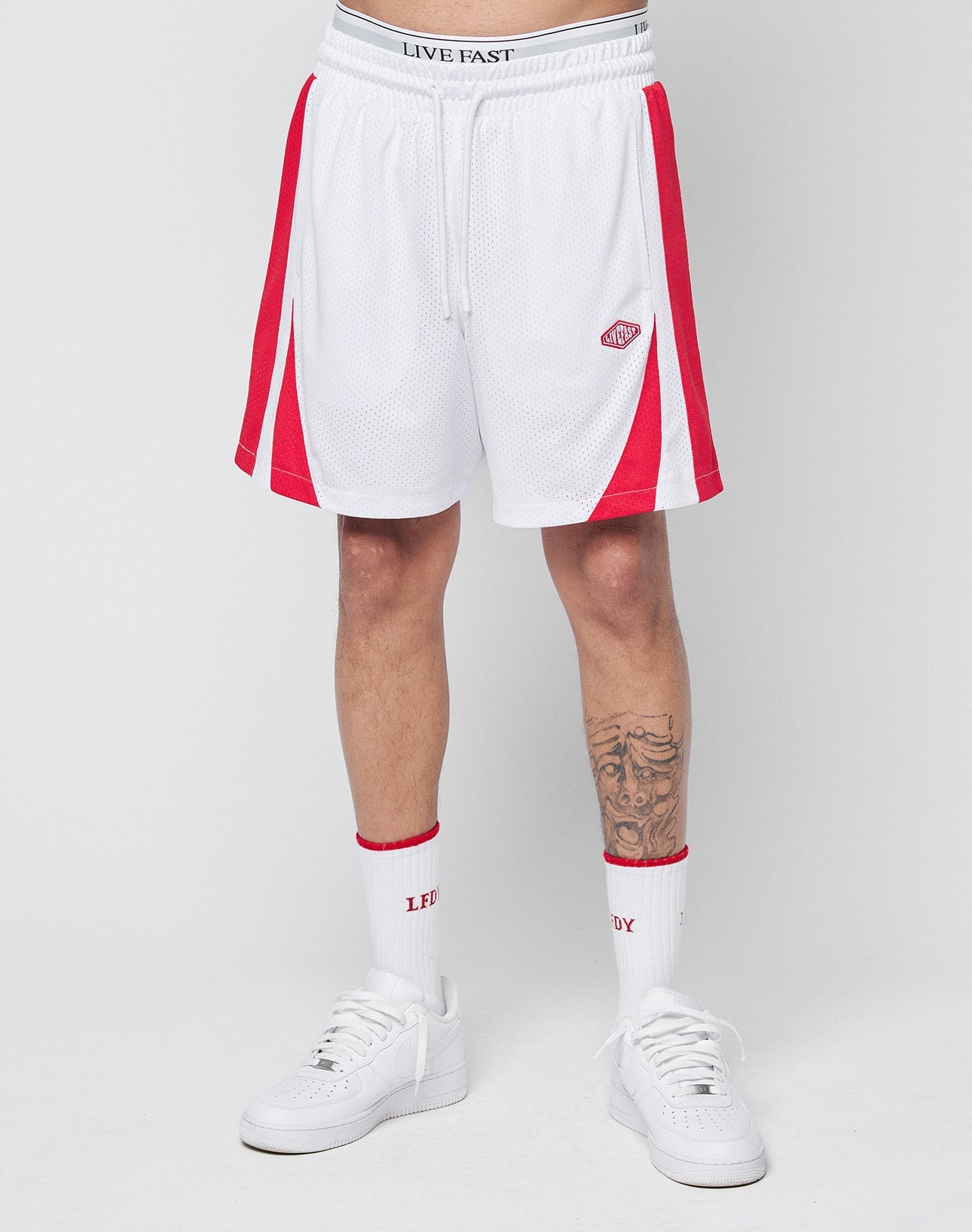 A person is seen from the waist down wearing LIVE FAST DIE YOUNG Striped Mesh Shorts in white with red stripes, a logo patch on the left leg, and LIVE FAST on the waistband, styled with white LFLUX socks and white sneakers.