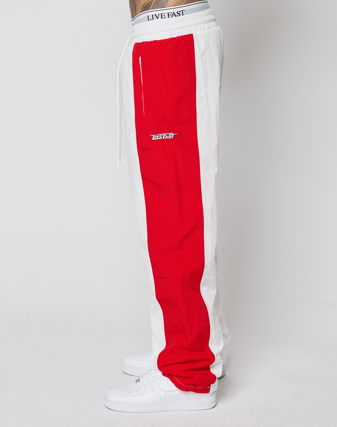 A person wears wide-fit white sneakers and LIVE FAST DIE YOUNGs LFDY Trackpants 3000—white track pants featuring a bold red stripe and LIVE FAST on the elastic waistband—standing against a plain white background.