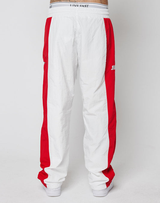 A person wears LIVE FAST DIE YOUNGs LFDY Trackpants 3000—wide fit, white with bold red side panels, an elastic waistband reading LIVE FAST, and a small left leg logo—styled with white sneakers against a plain background.