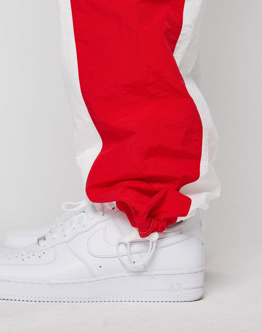 A close-up of a person wearing white Nike Air Force 1 sneakers and red and white LIVE FAST DIE YOUNG LFDY Trackpants 3000 with elastic cuffs and Wide Fit, standing against a plain white background.