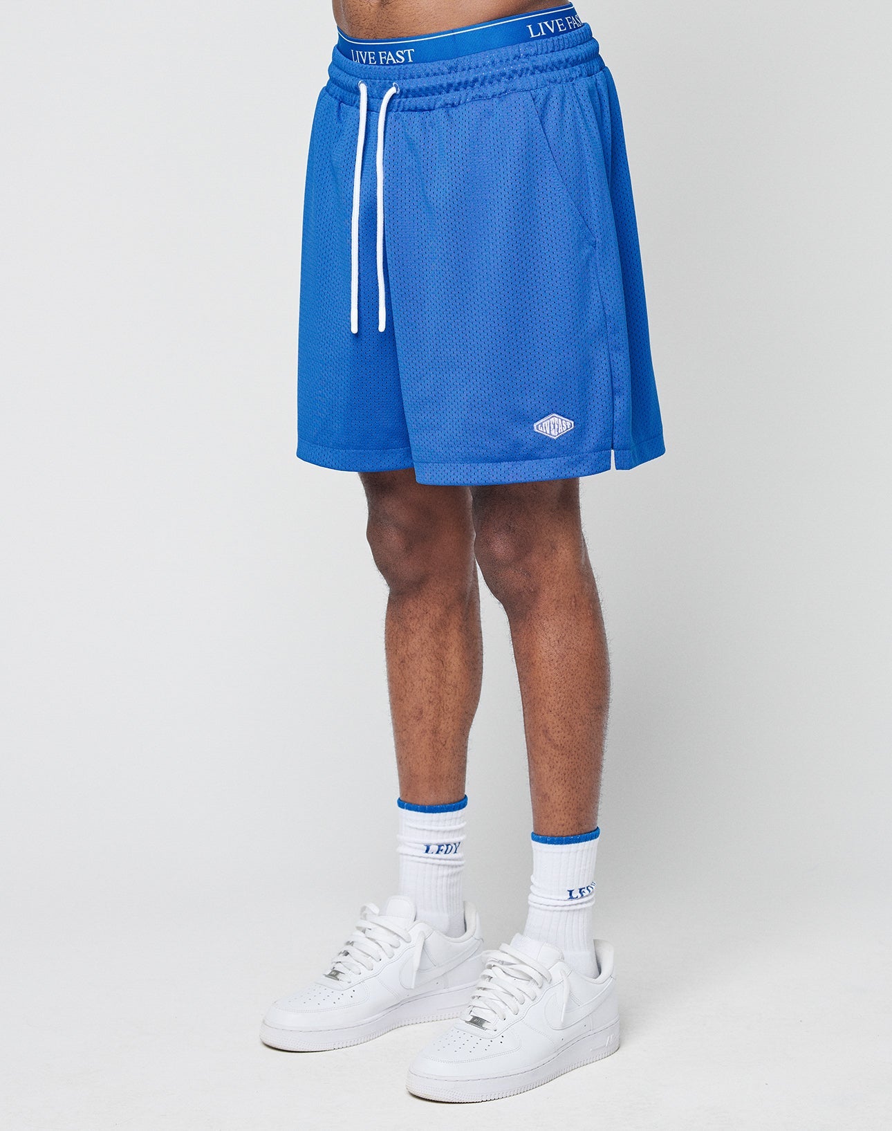 A person wears blue Basic Mesh Shorts by LIVE FAST DIE YOUNG with an elastic waistband and drawstring featuring LIVE FAST, paired with white socks with blue trim and white sneakers, standing against a plain white background. | model
