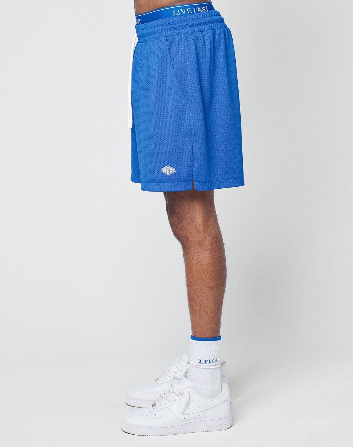 A person stands sideways in LIVE FAST DIE YOUNG Basic Mesh Shorts (blue, regular fit, elastic waist), white LIVE FAST socks, and white sneakers. Only the lower torso and legs are shown against a plain white background.