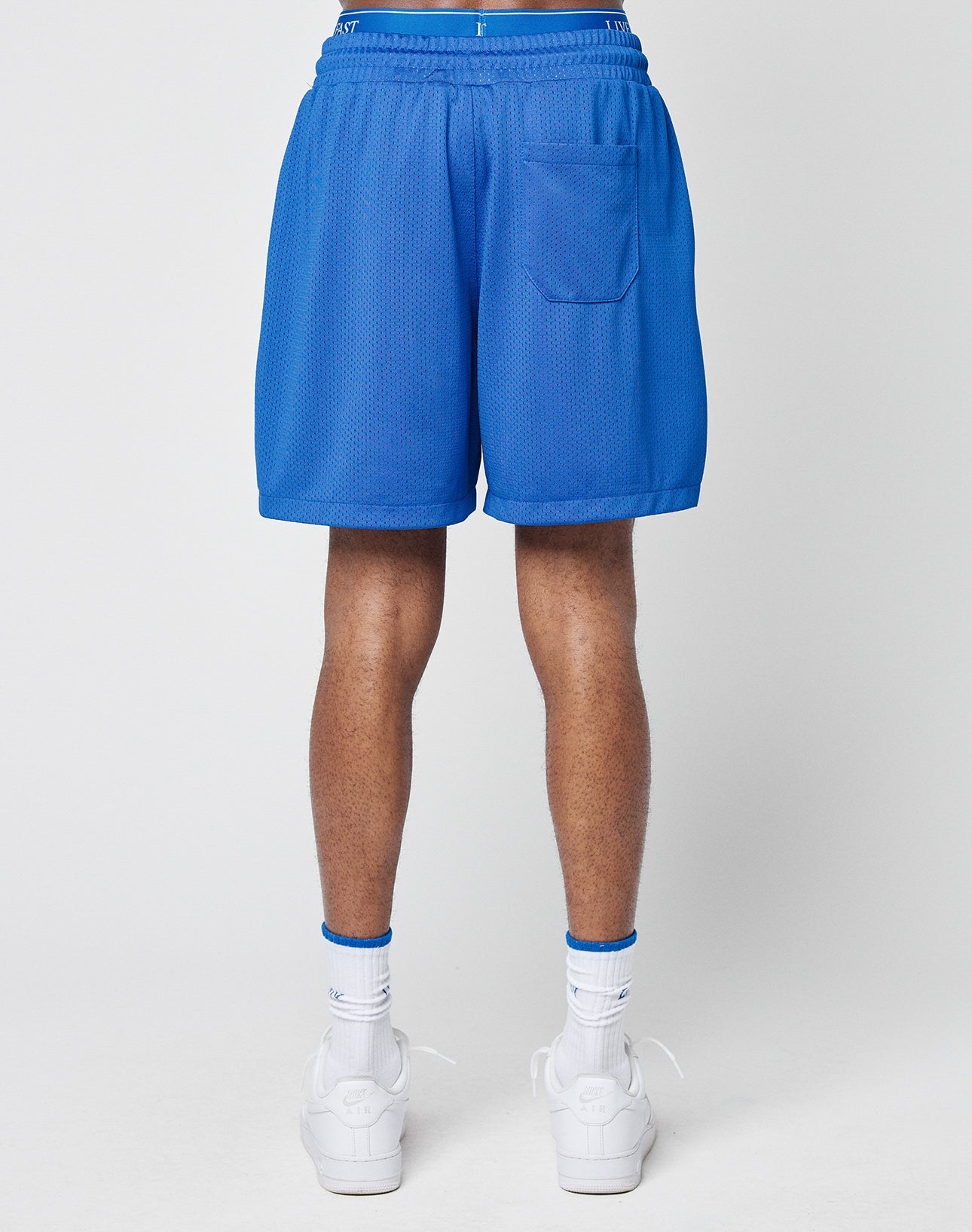 A person stands facing away, wearing LIVE FAST DIE YOUNG Basic Mesh Shorts in bright blue with an elastic waistband and back pocket, white crew socks with blue stripes, and white sneakers against a plain light background.